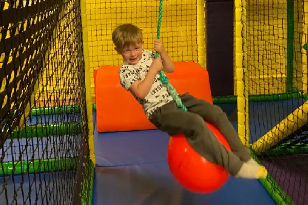 Young child playing on a red ball in a colourful soft play structure with netting.