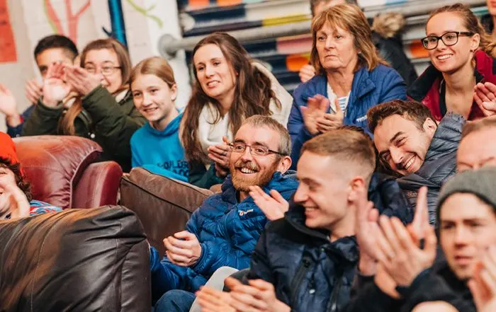 A group of children and adults laughing and enjoying time together in a casual indoor setting with colourful artwork visible in the background.