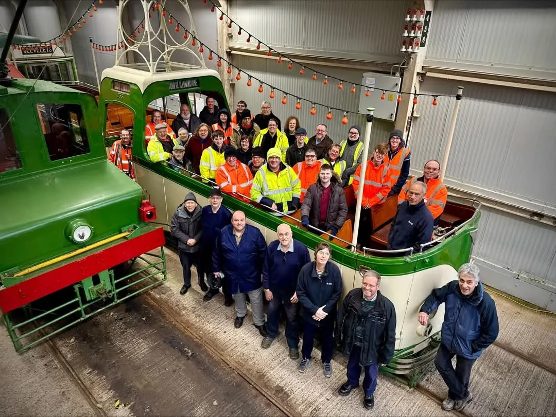 Large group of visitors and staff posing with a vintage green and cream tram inside a heritage railway depot.