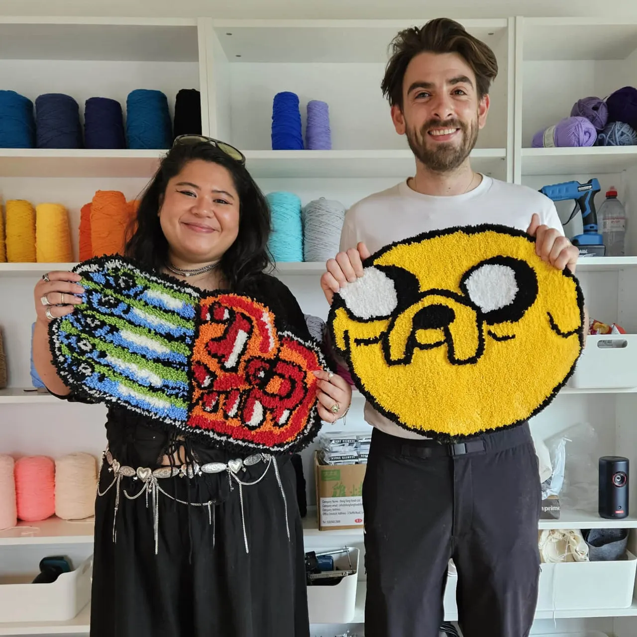 Two people displaying colourful tufted rugs they've made in a craft studio, with shelves of yarn behind them.