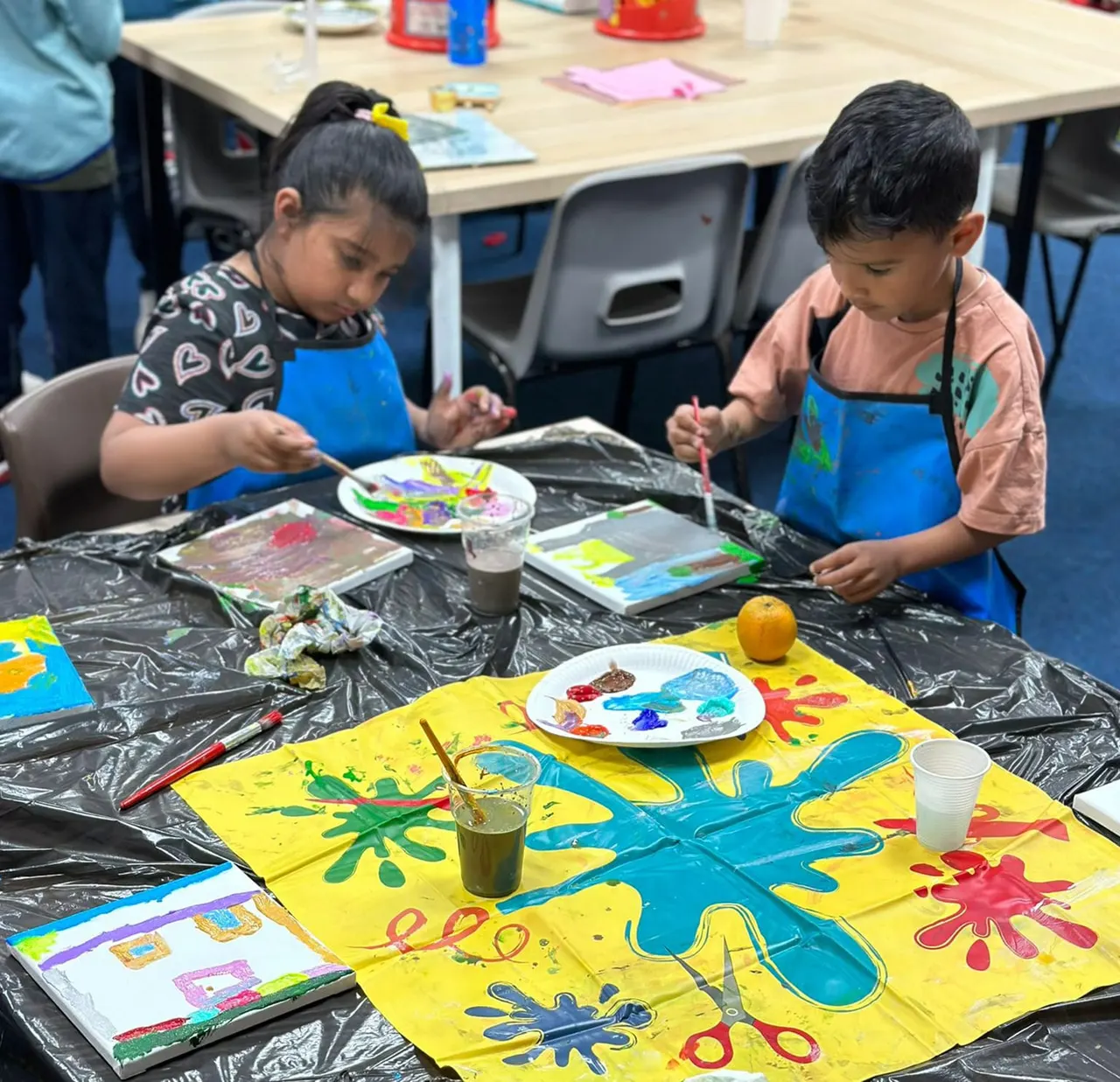 Two young children wearing blue aprons paint at a table covered with colourful artwork and art supplies during a creative activity session.