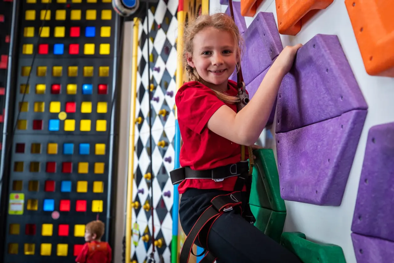 Young child in red shirt climbing coloured foam holds on an indoor climbing wall, wearing a safety harness.
