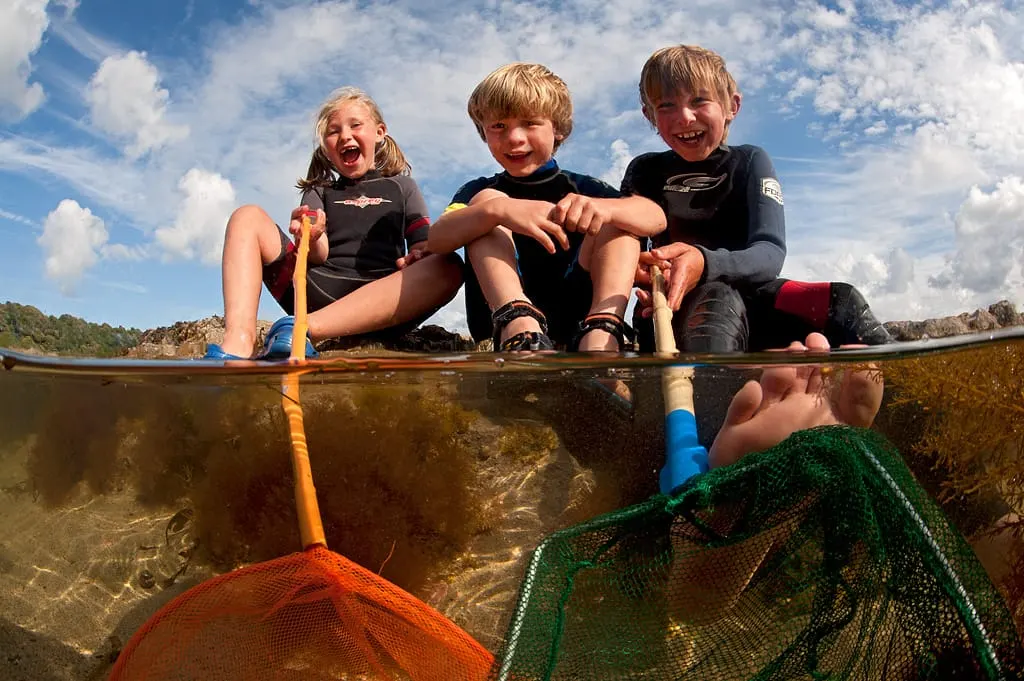 Three children sit in a boat holding fishing nets above a brown waterway, smiling at the camera.