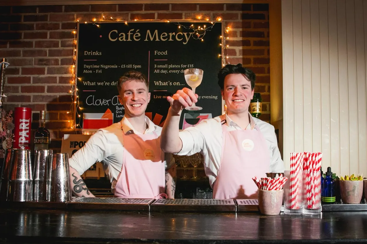 Two smiling bartenders in pink aprons stand behind a counter holding a cocktail glass at Café Mercy.