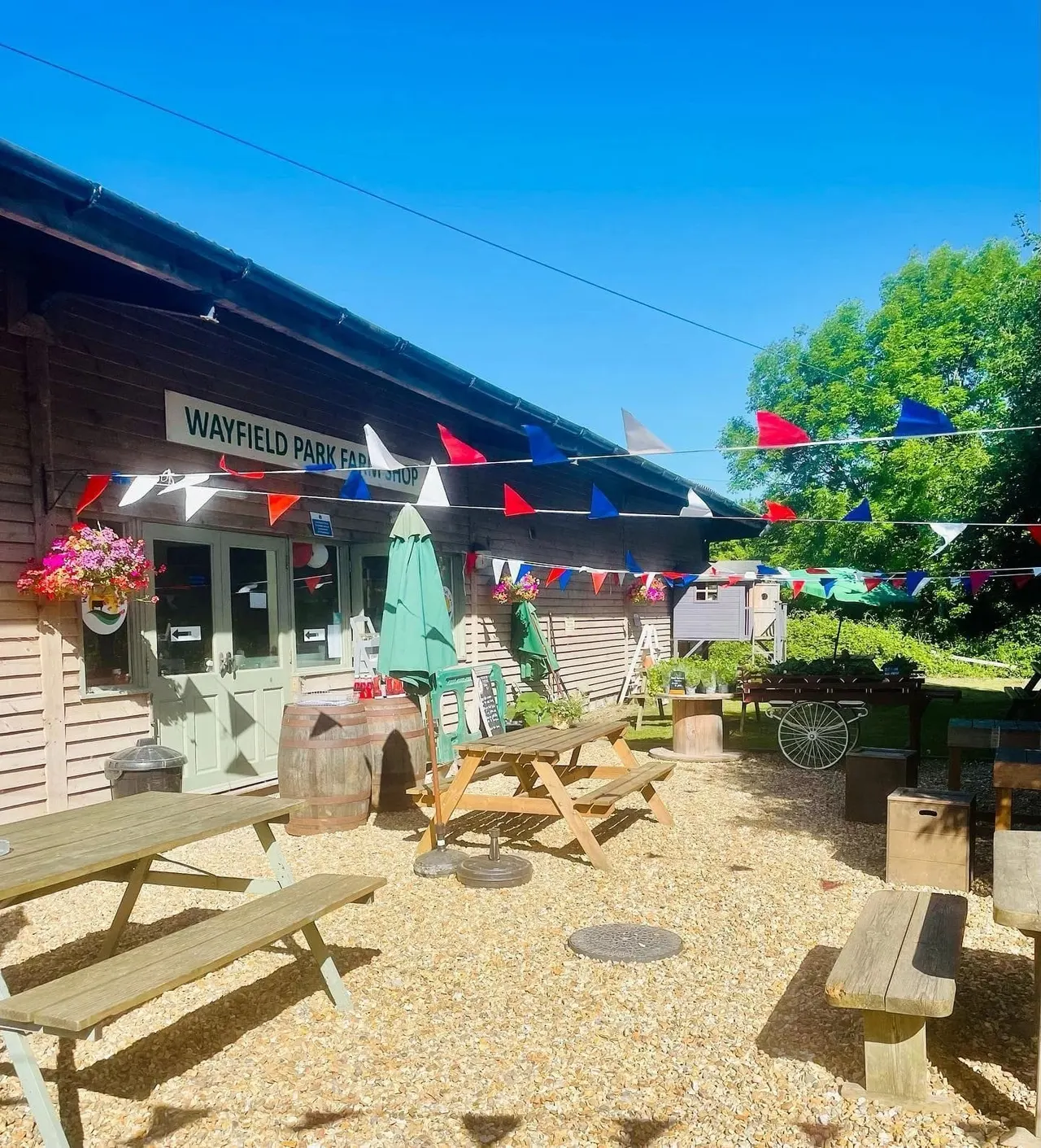 Outdoor seating area at a farm venue with picnic tables, bunting, potted flowers, and a green building under blue sky.