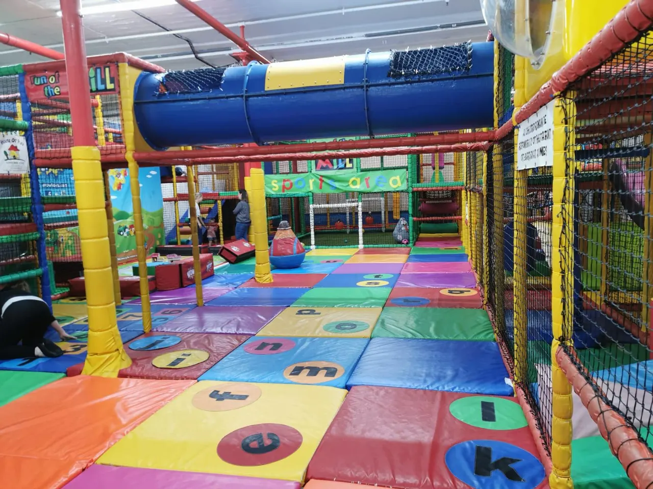 Colourful indoor soft play area with padded floor, inflatable tunnel, climbing structures and numbered play mats, children playing throughout.