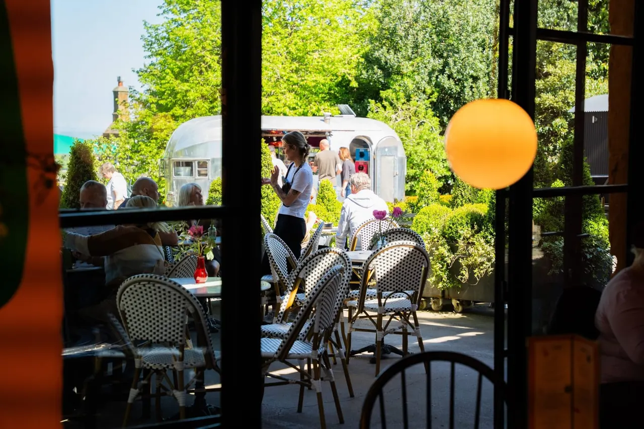 View through windows of an indoor café onto a sunny outdoor terrace with vintage caravans, seated guests, and lush green surroundings.