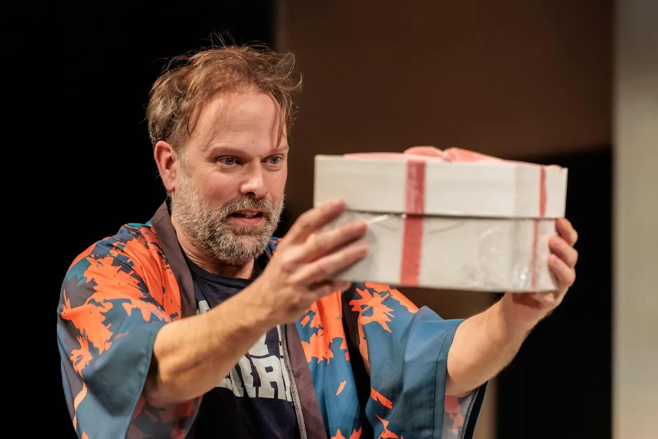 Man in colourful jacket holding a wrapped gift box whilst speaking on stage.