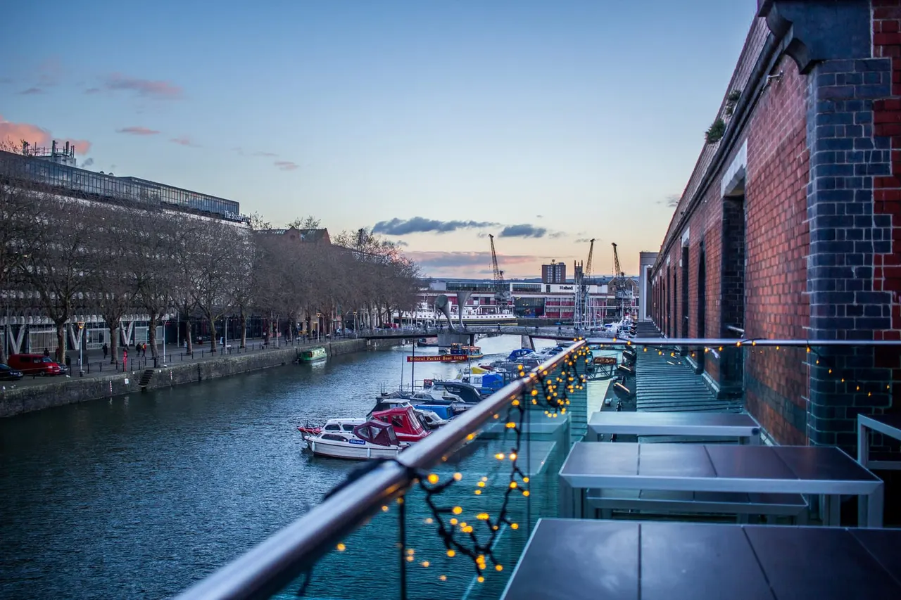 Dusk view of a waterfront dock with moored boats, brick buildings, and festive lighting along a railing overlooking the water.