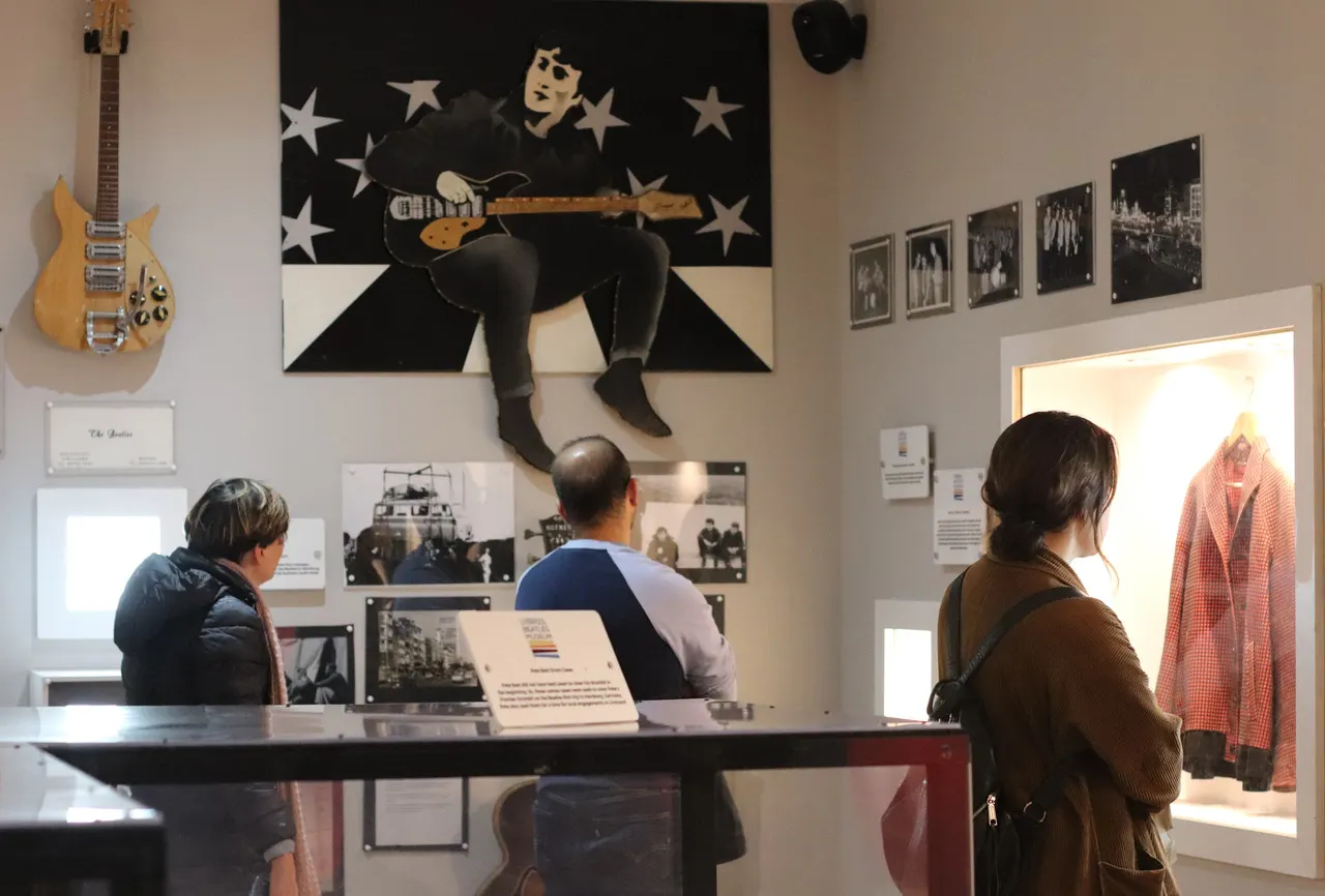 Visitors viewing a music museum exhibition with a large portrait artwork of a musician with a guitar, vintage guitars and photographs on display walls.