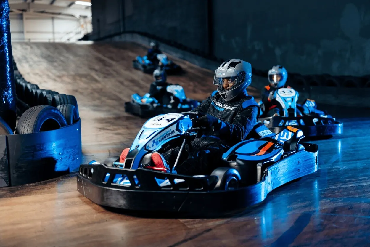 Indoor go-kart racer in blue and black suit navigating track with other karts and barriers visible.