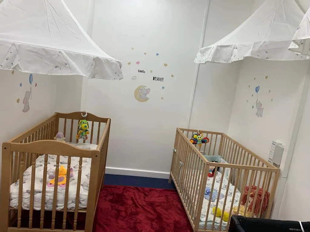 Two wooden baby cots with toys inside in a soft play area decorated with white canopies and moon and stars wall art.