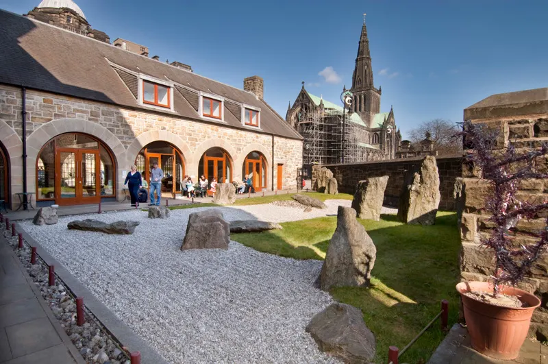 Stone museum building with arched entrances next to historic ruins and cathedral in background, visitors outside on sunny day.