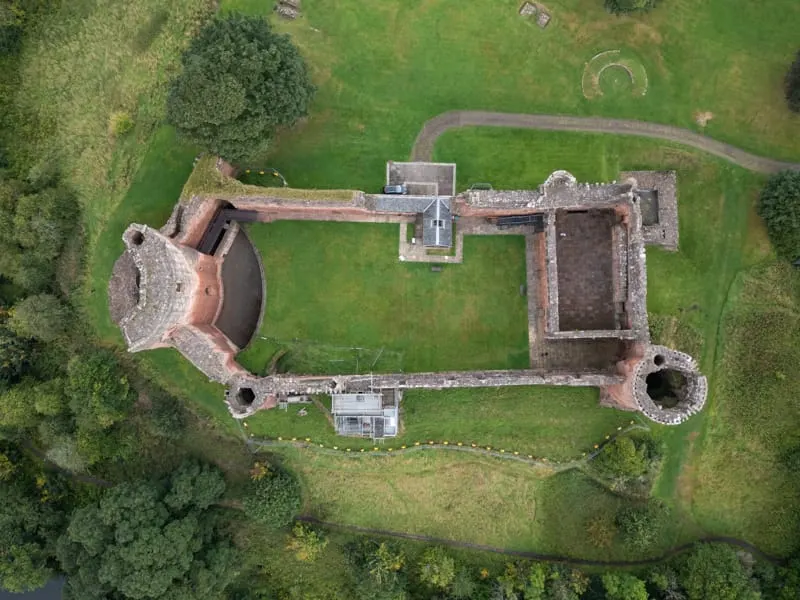 Aerial view of a ruined castle with circular towers and connecting walls surrounded by green lawns and woodland.