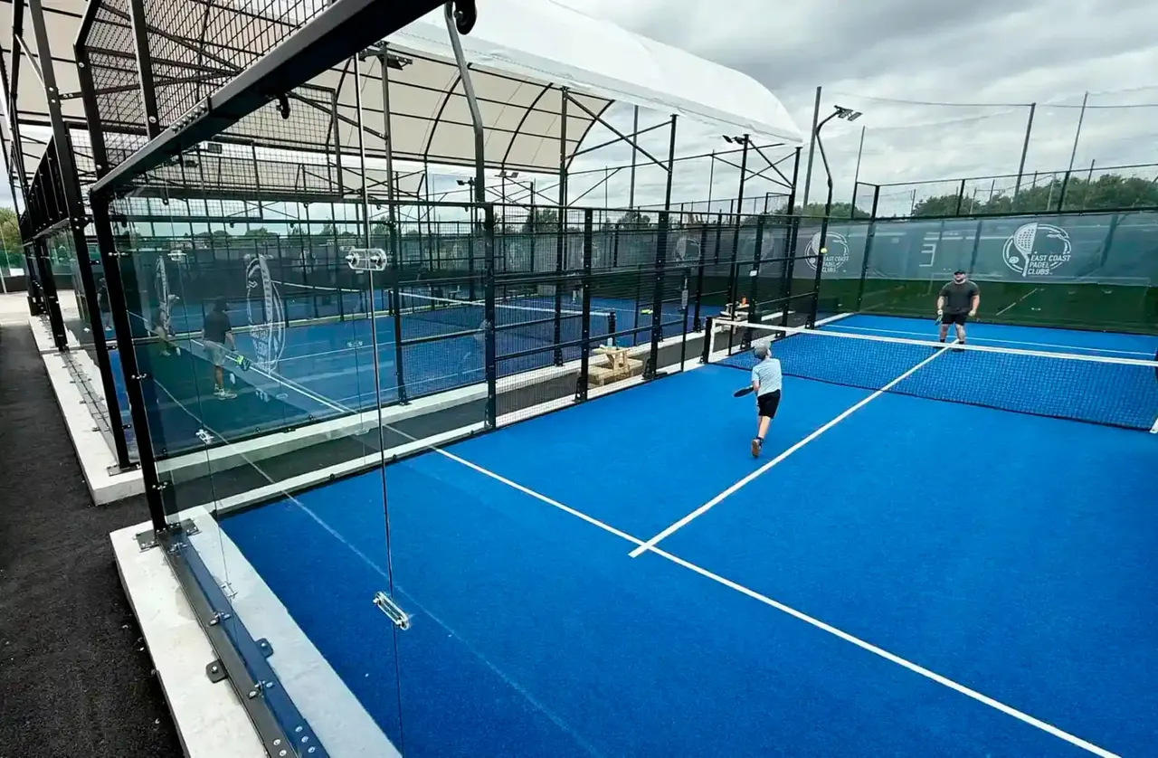 Multiple players on blue padel tennis courts under a white canopy roof at an outdoor sports facility.