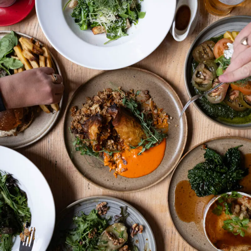 Multiple plated dishes shared at a dining table including meat with red sauce, greens, fries, and salads, with diners' hands visible.