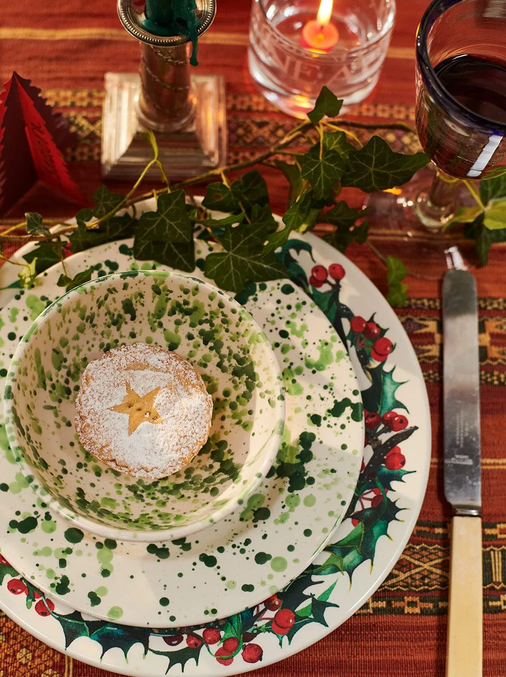 Festive table setting with a star-shaped pastry dusted with icing on holly-patterned plates, wine glasses and ivy decoration.