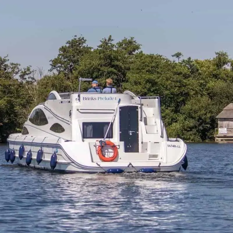 White motor cruiser moored on a river with two people visible on the roof, trees lining the waterside behind.