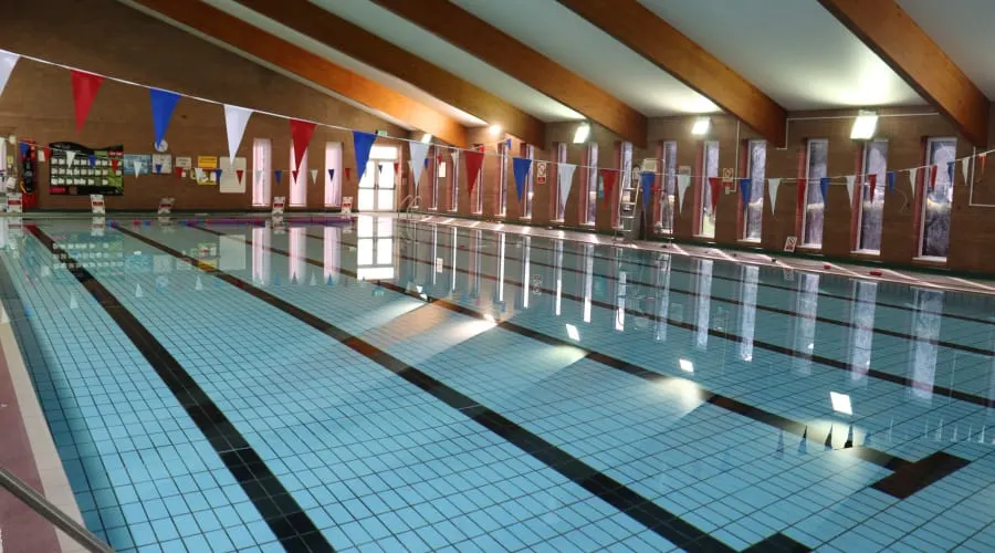Indoor swimming pool with multiple lanes, wooden beams, bunting, and spectator gallery with glass barriers.