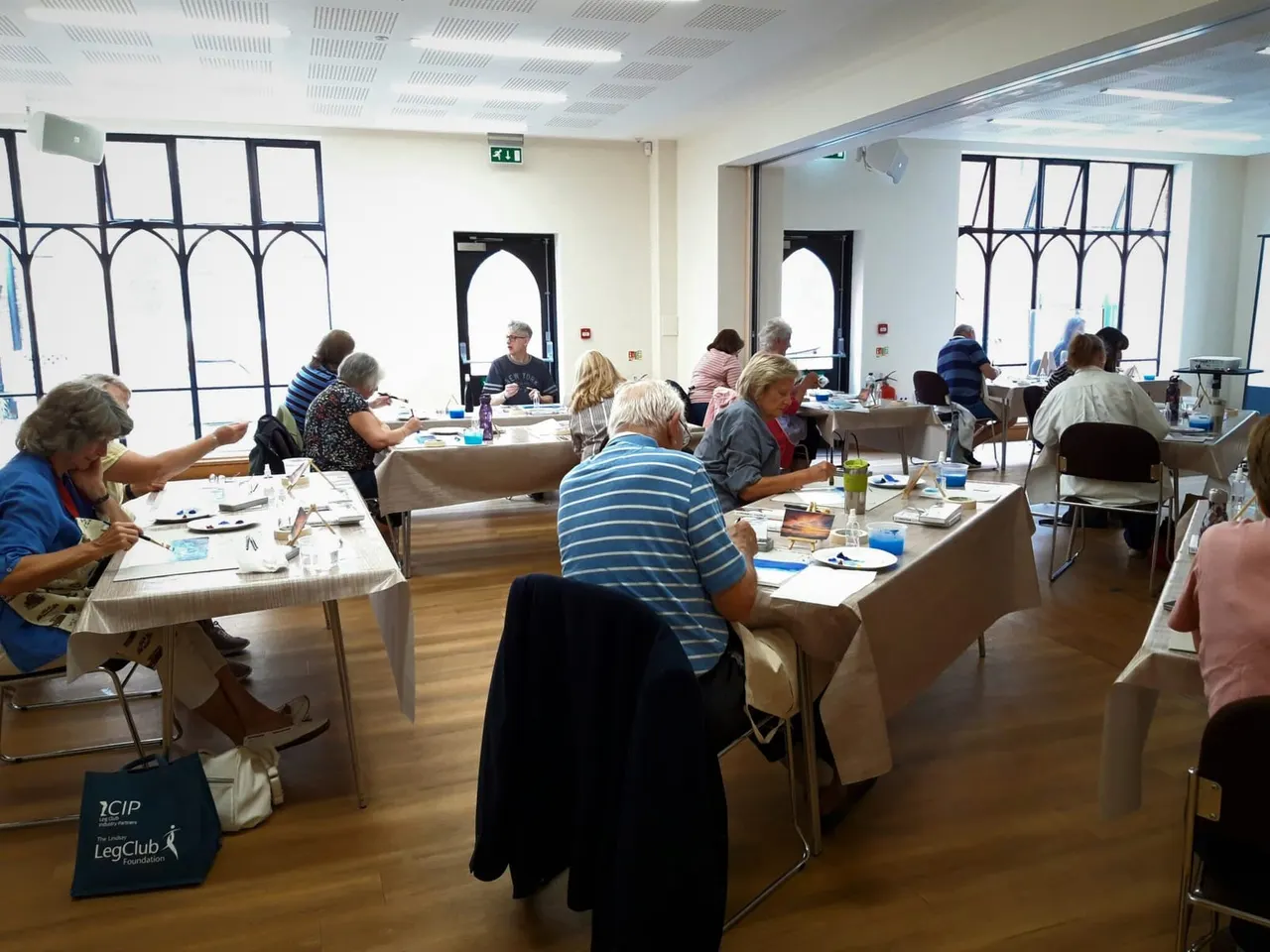 Adults seated at tables during an art class in a bright, modern studio with large windows.