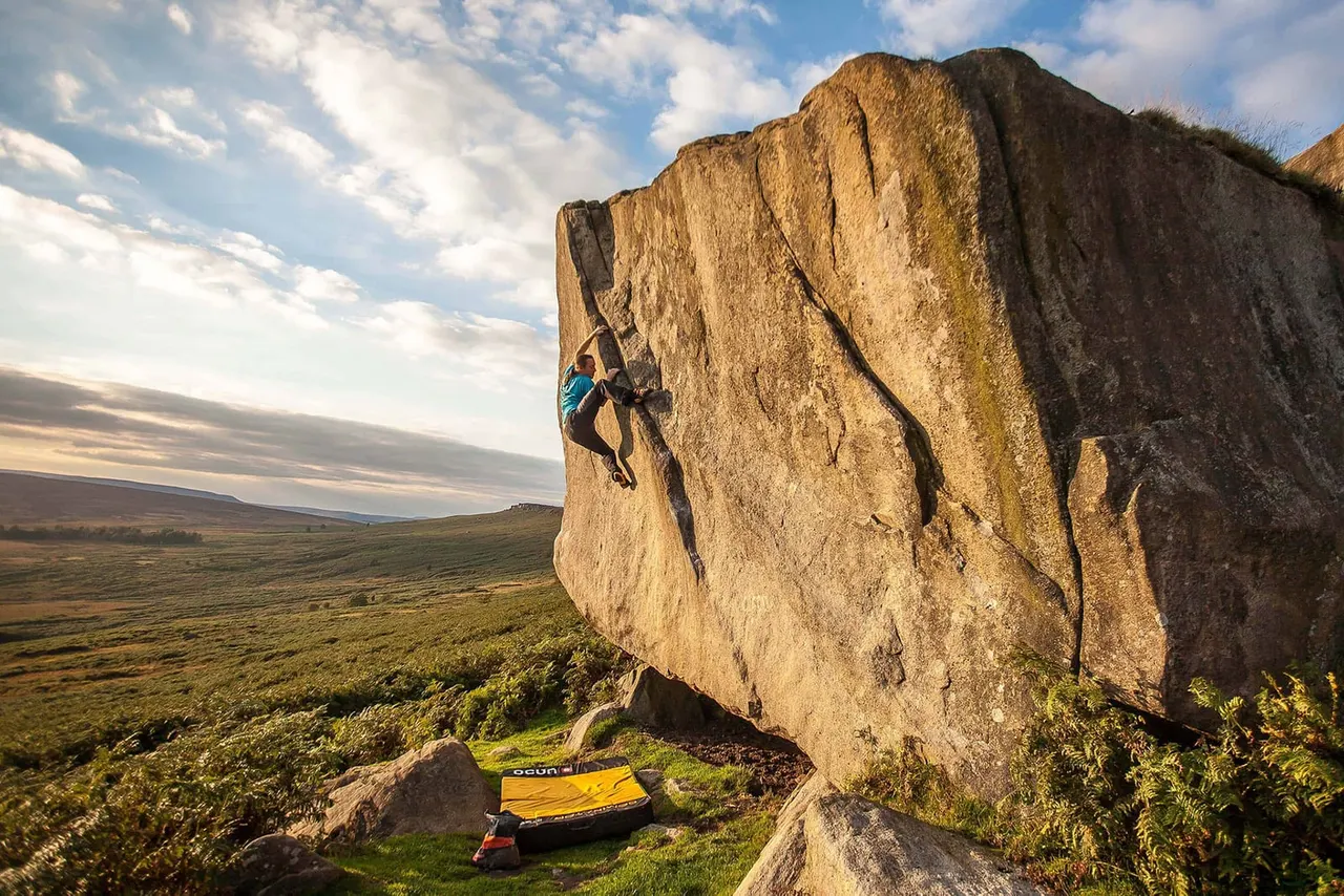 A person rock climbing on a large natural boulder outcrop in open moorland with climbing equipment laid on the grass below.