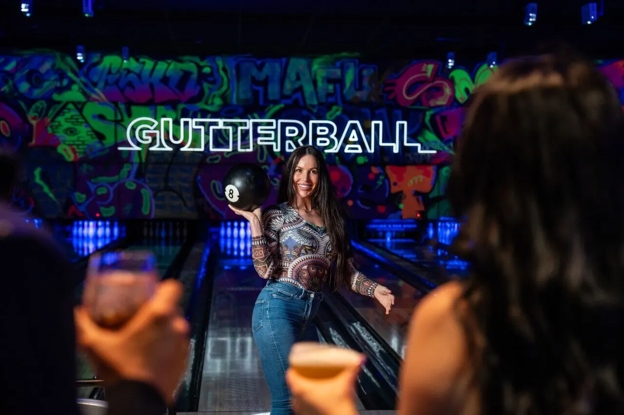 Woman holding a number 8 ball on a neon-lit gutterball lane with colourful graffiti-style graphics on the wall behind her.