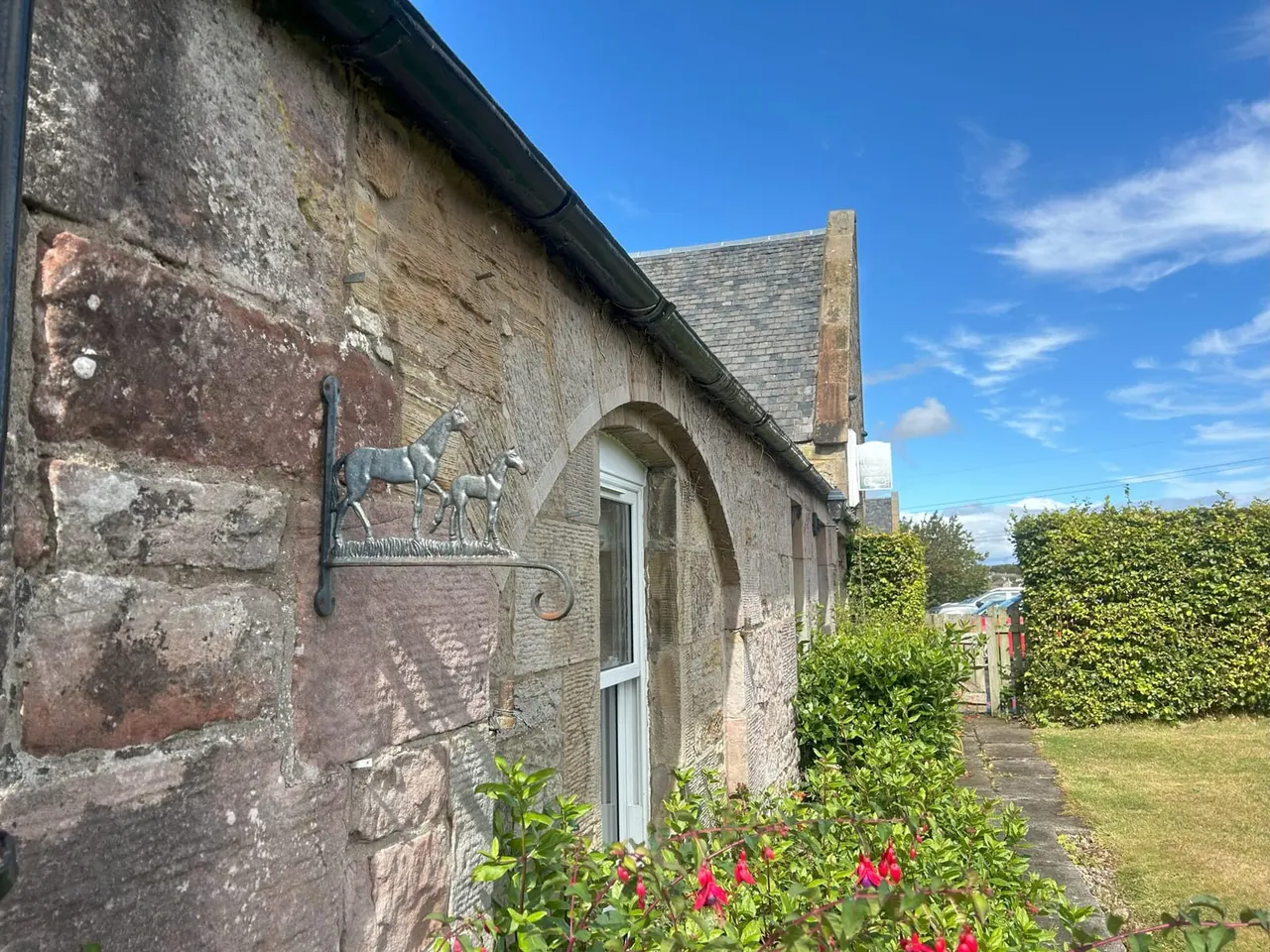 Historic stone cottage with decorative horse weathervane, arched doorway and slate roof, surrounded by gardens and hedging on a sunny day.