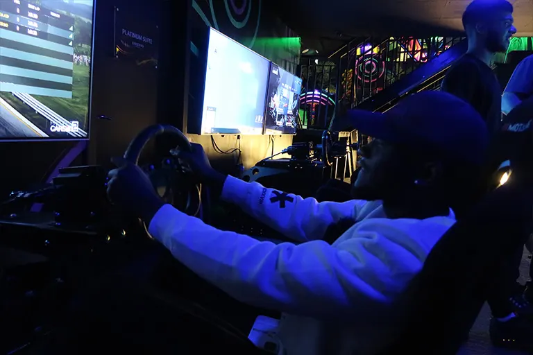 Person in racing simulator cockpit with blue lighting and screens in entertainment arcade.