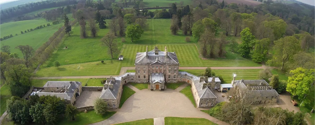 Aerial view of a Georgian mansion with formal grounds, flanked by stone estate buildings, surrounded by rolling countryside and tree-lined fields.
