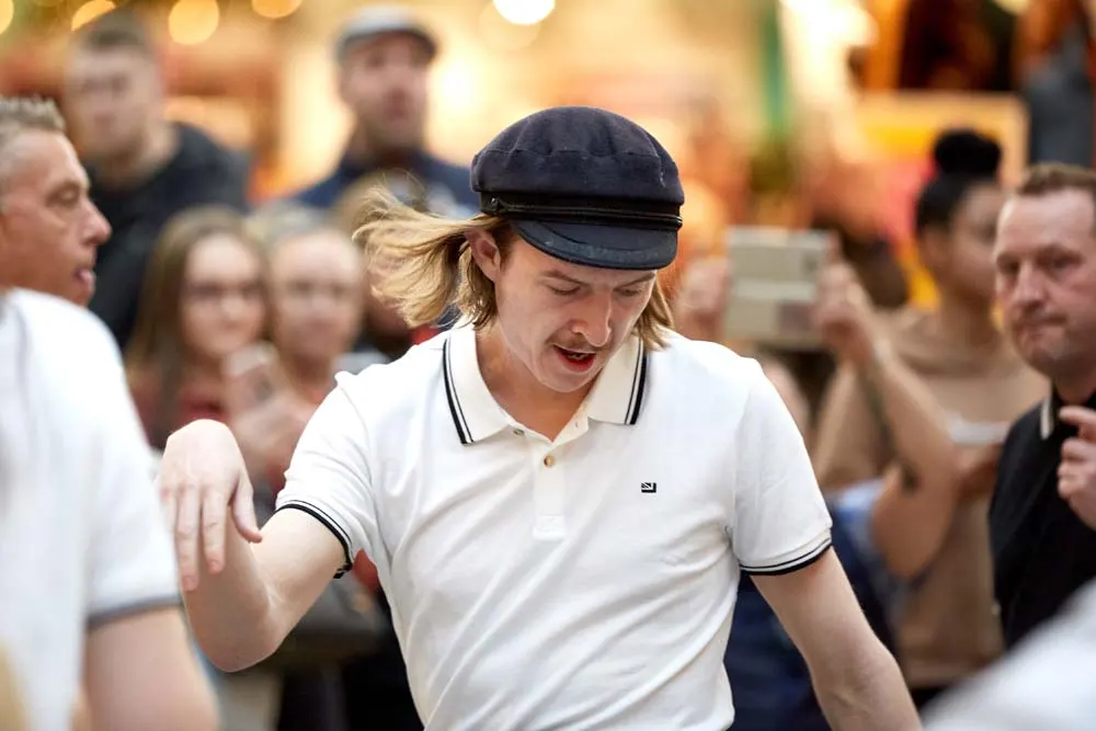 Person in white polo shirt and black cap dancing enthusiastically in front of a crowd at an indoor venue.