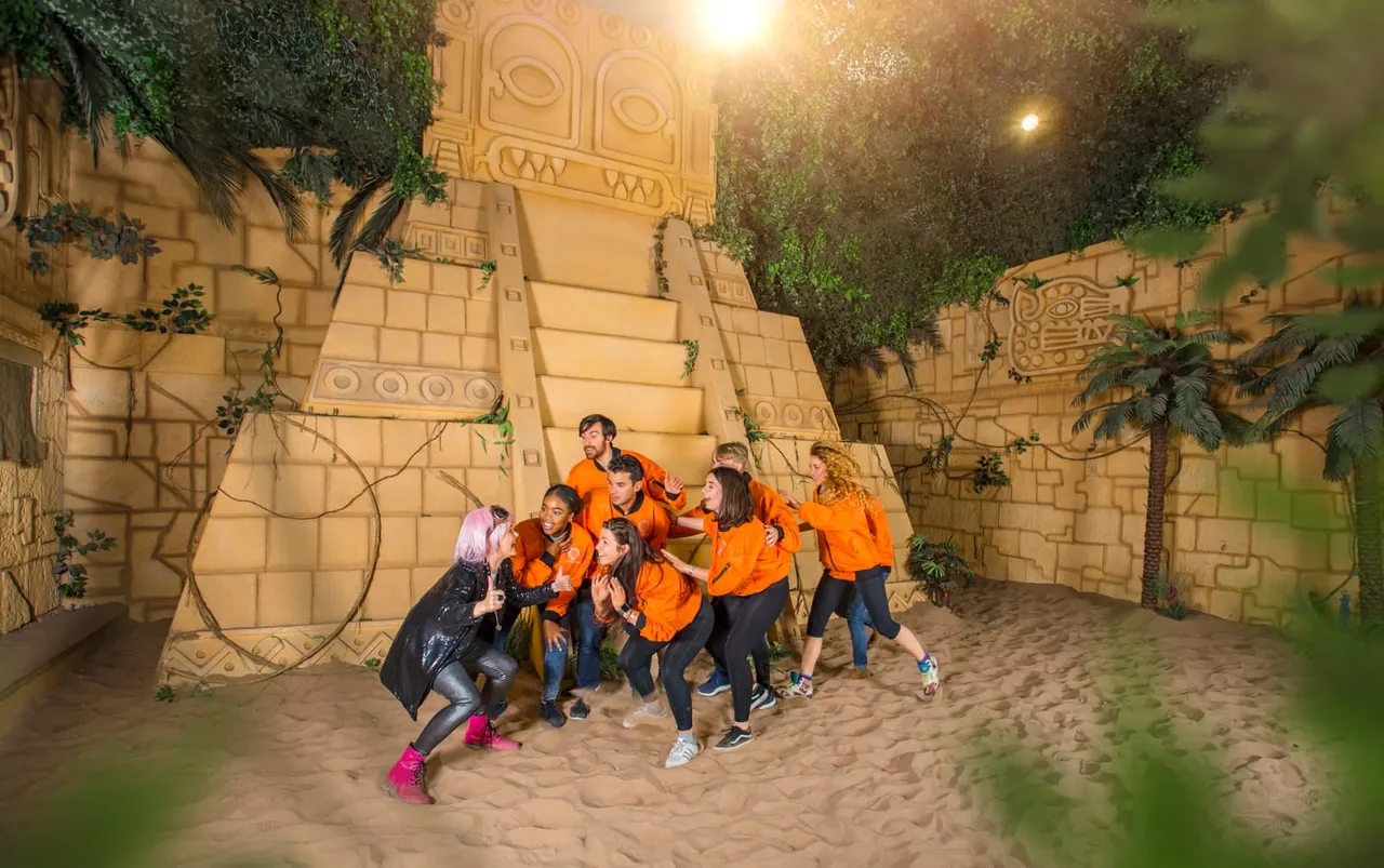 Group of children in orange shirts playing together in a sandy arena with tall decorated stone walls and climbing structures.