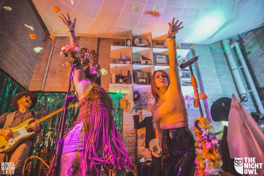 Live band performing on stage with colourful lighting, featuring musicians with guitars and singers with raised arms in an energetic indoor venue.