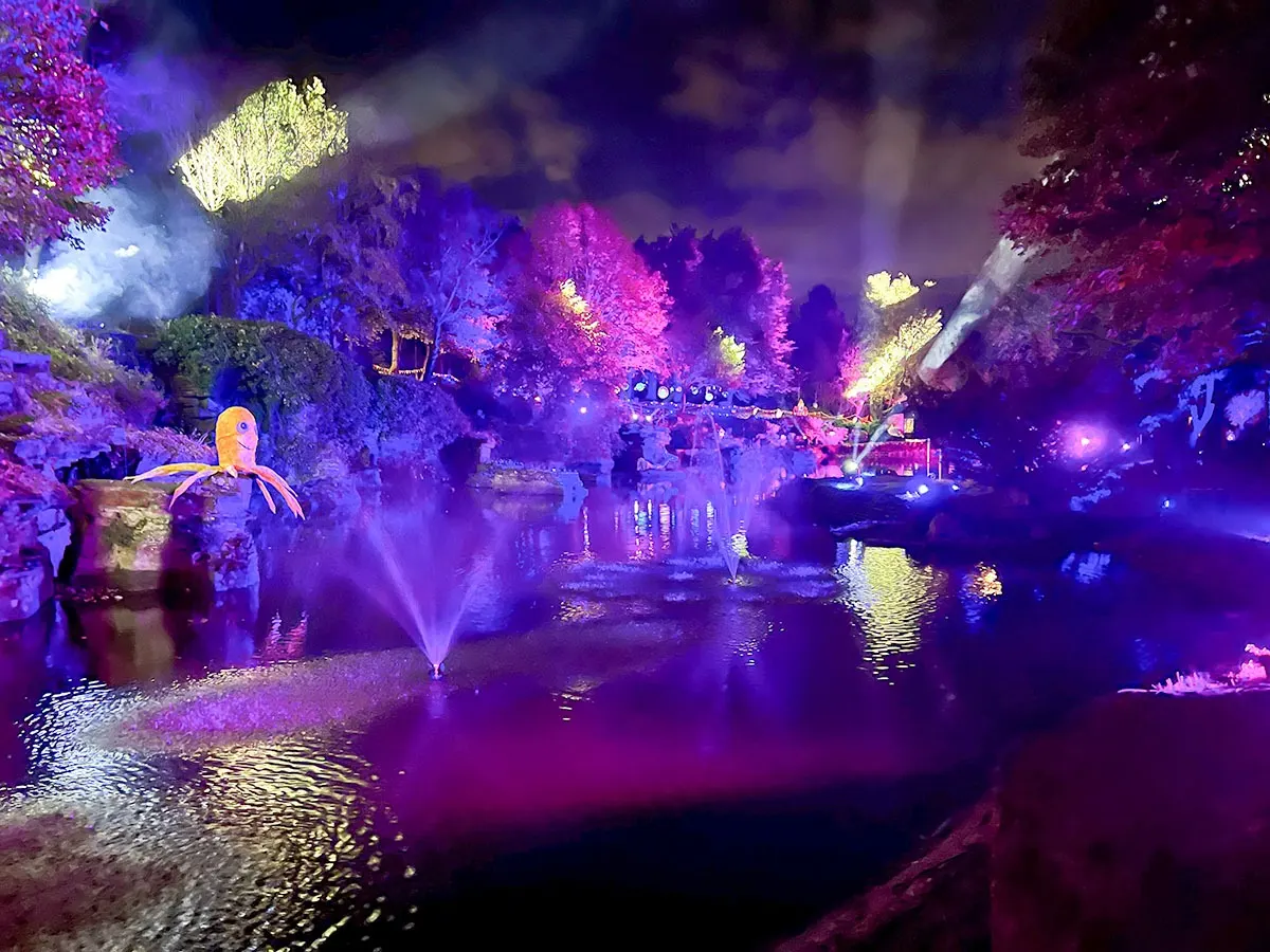 Night-time outdoor garden display with purple and yellow illuminated trees, water features, fountains, and decorative sculptures around a pond.