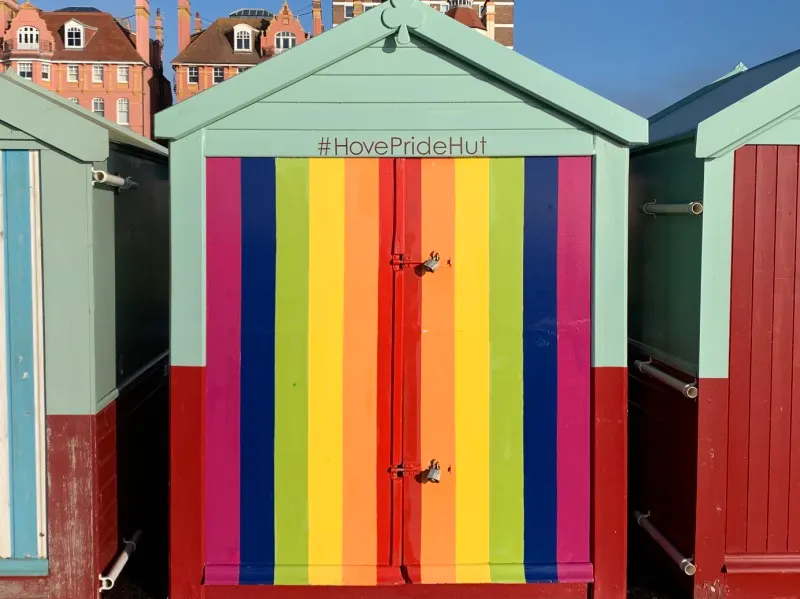A rainbow-striped beach hut with #HovePrideHut hashtag, flanked by other pastel-coloured huts and Victorian buildings behind.