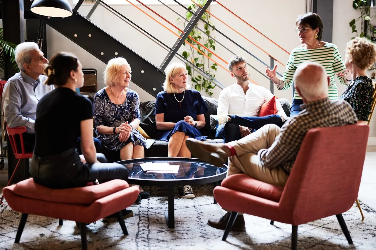A group of adults seated in a circle on red chairs in a modern lounge space, engaged in conversation around a central table.