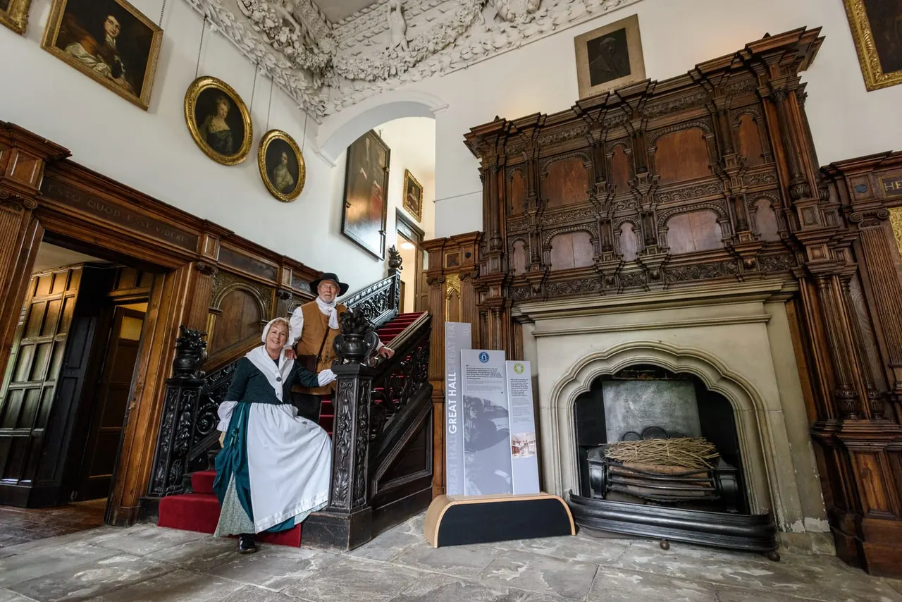 Two costumed interpreters stand on an ornate wooden staircase in a grand historic hall with decorative ceiling, portraits, and a large fireplace.