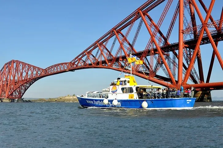 A blue and white passenger ferry passes beneath the iconic red cantilever bridge over the Firth of Forth.