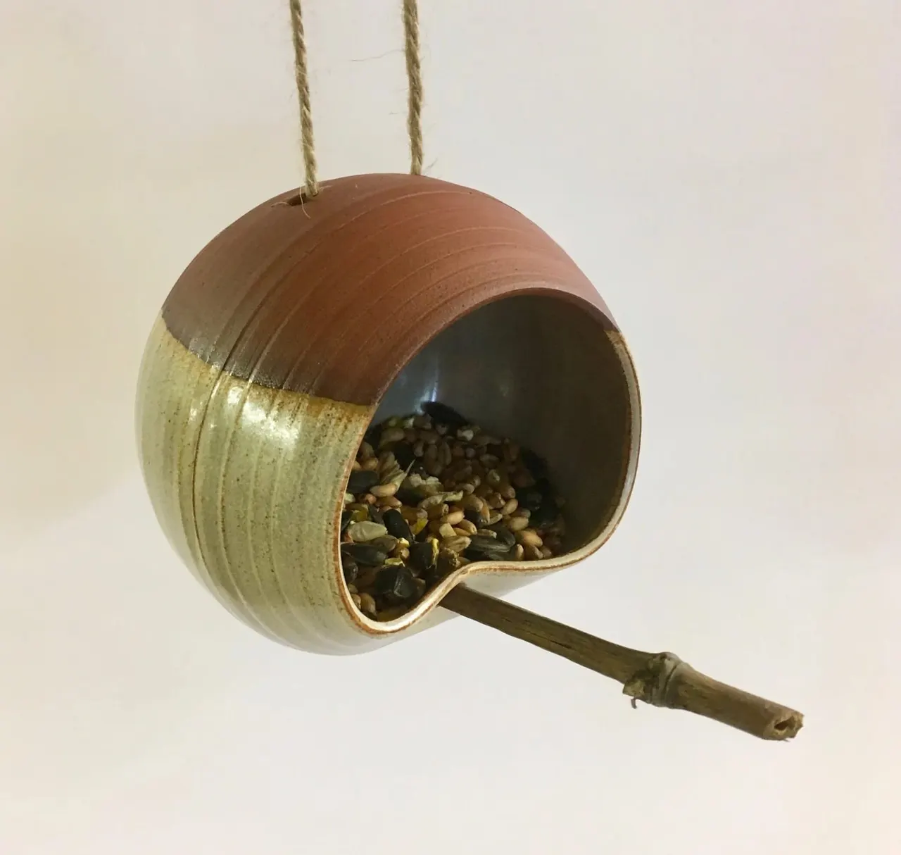 Ceramic bird feeder filled with seeds, hanging from rope against a white background.