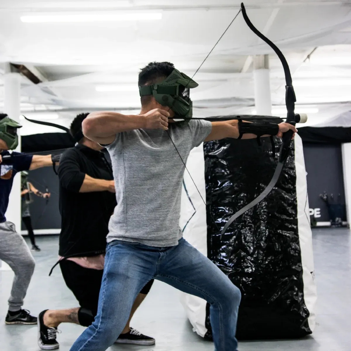 Person in protective mask drawing a compound bow at an indoor archery range with a target bag.