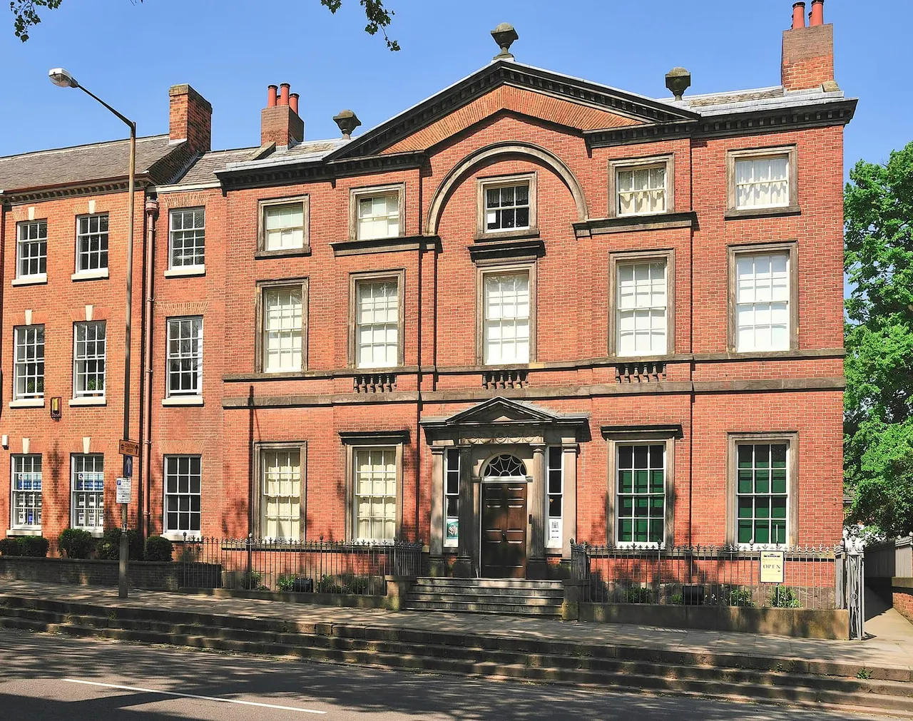 Exterior of a Georgian red-brick building with multiple storeys, sash windows, decorative stonework and chimneys, set behind iron railings on a sunny day.