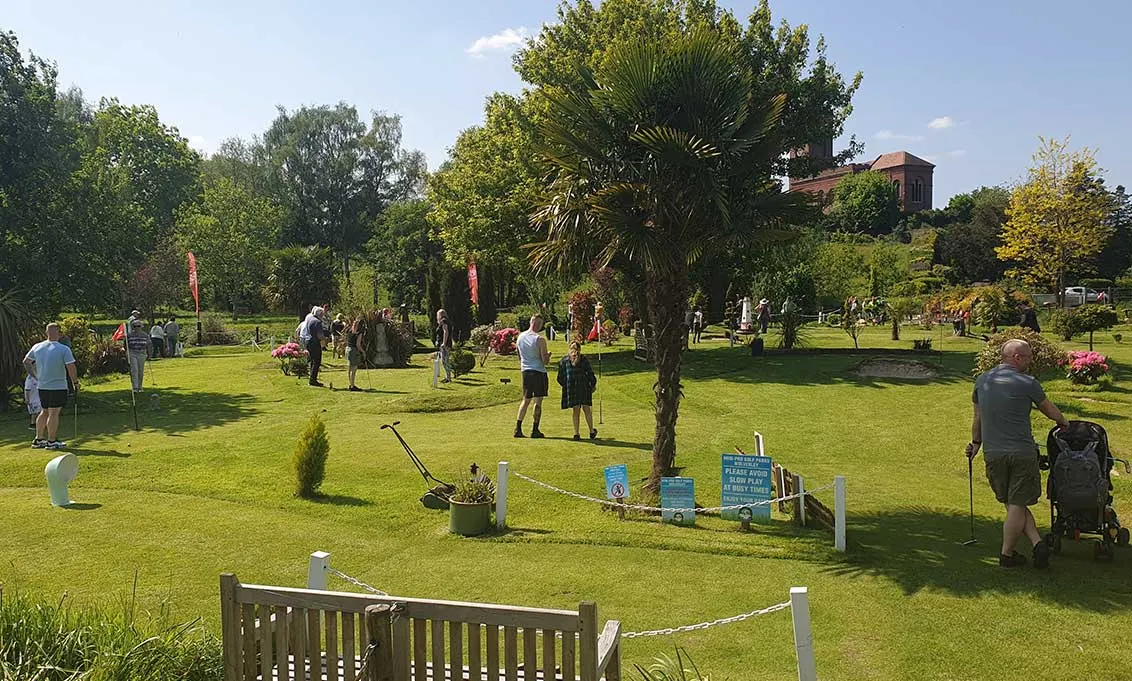 Visitors playing mini golf on a sunny day across a spacious green landscape with tropical plants and woodland.
