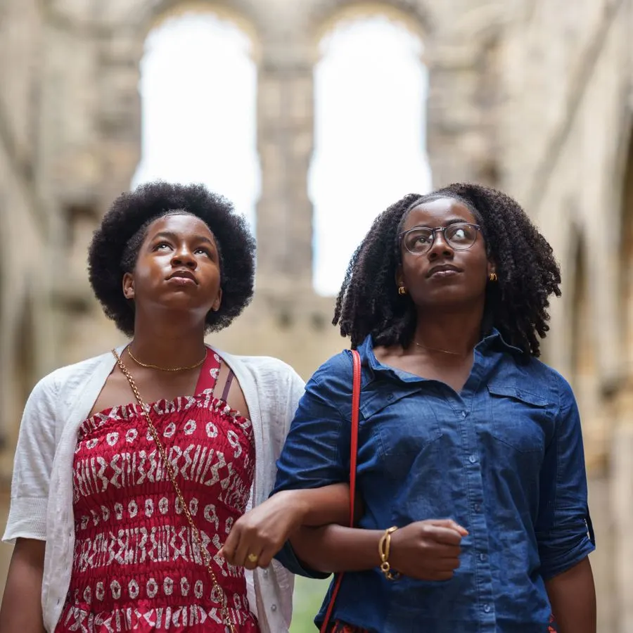 Two young women looking upward inside the ruins of a stone abbey with arched windows.
