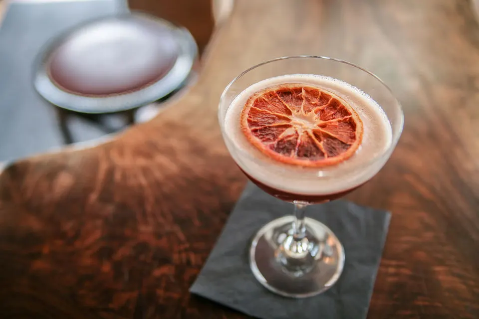 A grapefruit slice garnishes a creamy cocktail in a coupe glass on a dark coaster.