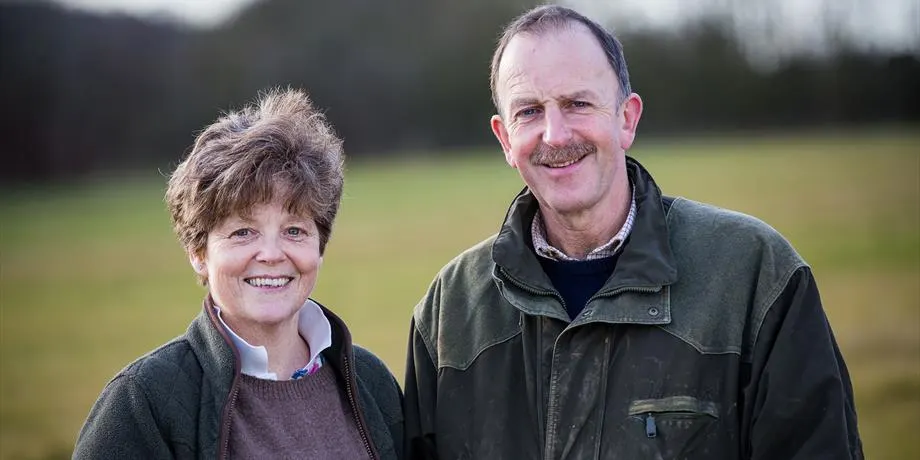 Two adults standing in a rural field, smiling at the camera with trees in the background.