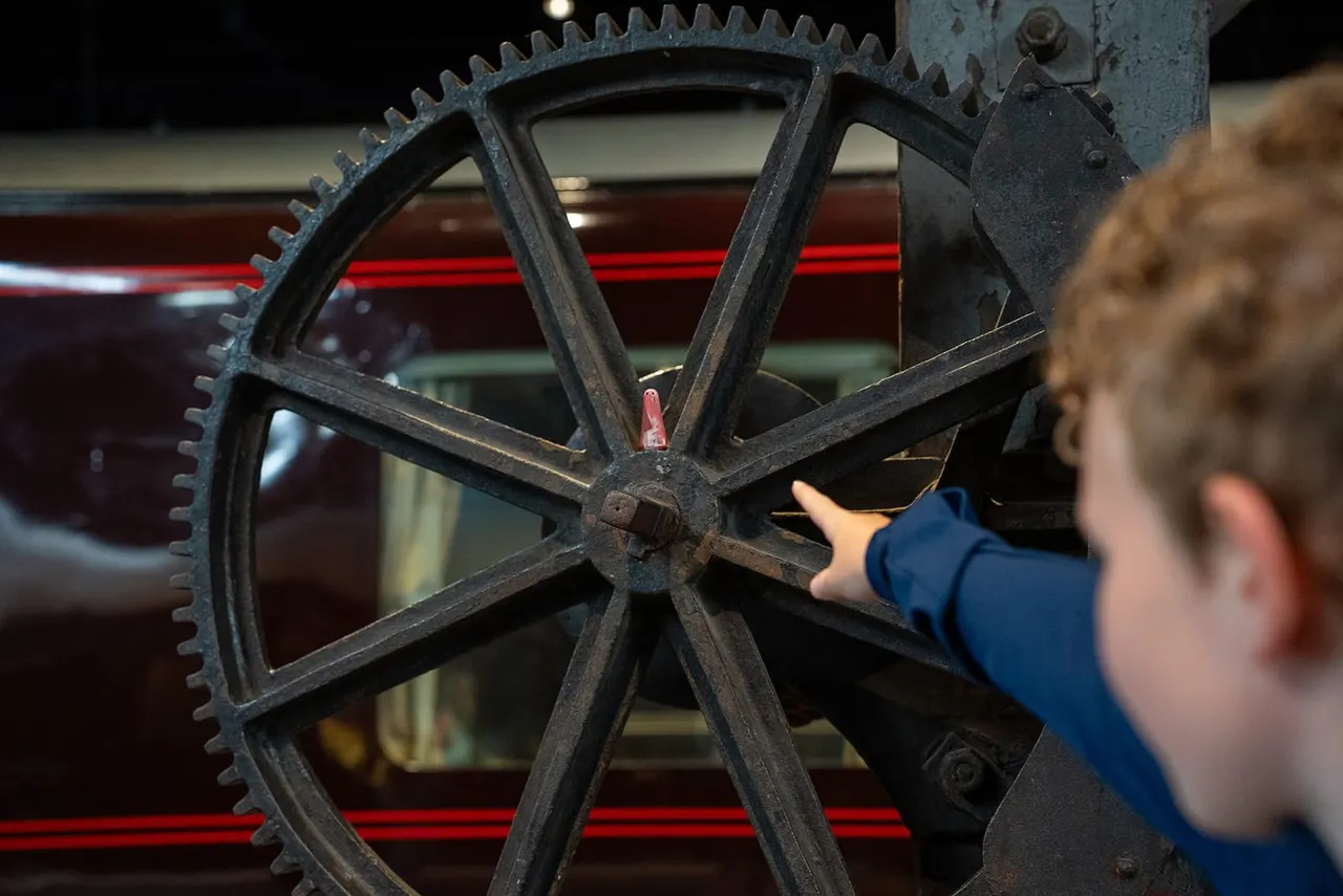 Child in blue jacket touching a large cast iron gear wheel from vintage industrial machinery.