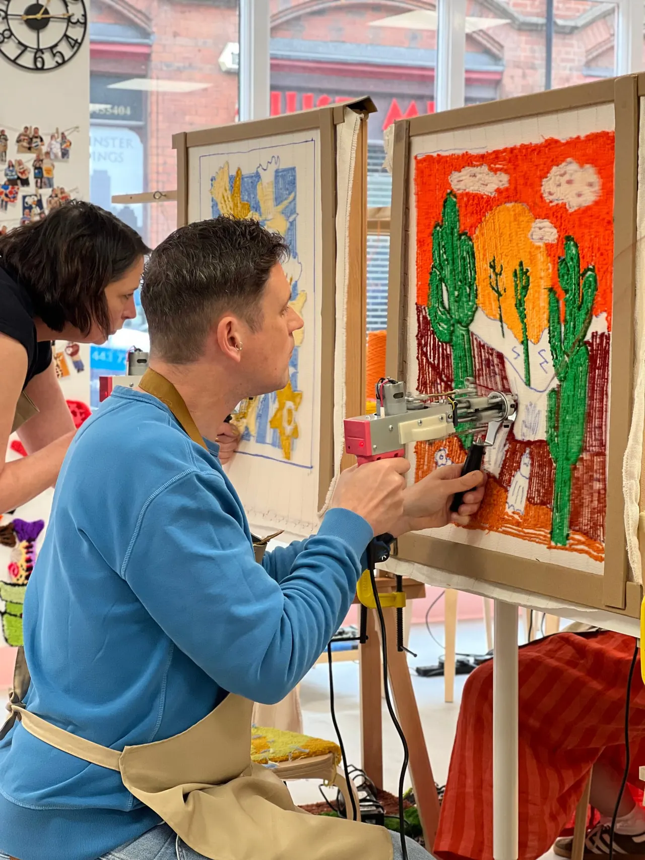 Two people operating a tufting gun on a framed textile artwork depicting a cactus against an orange sky.