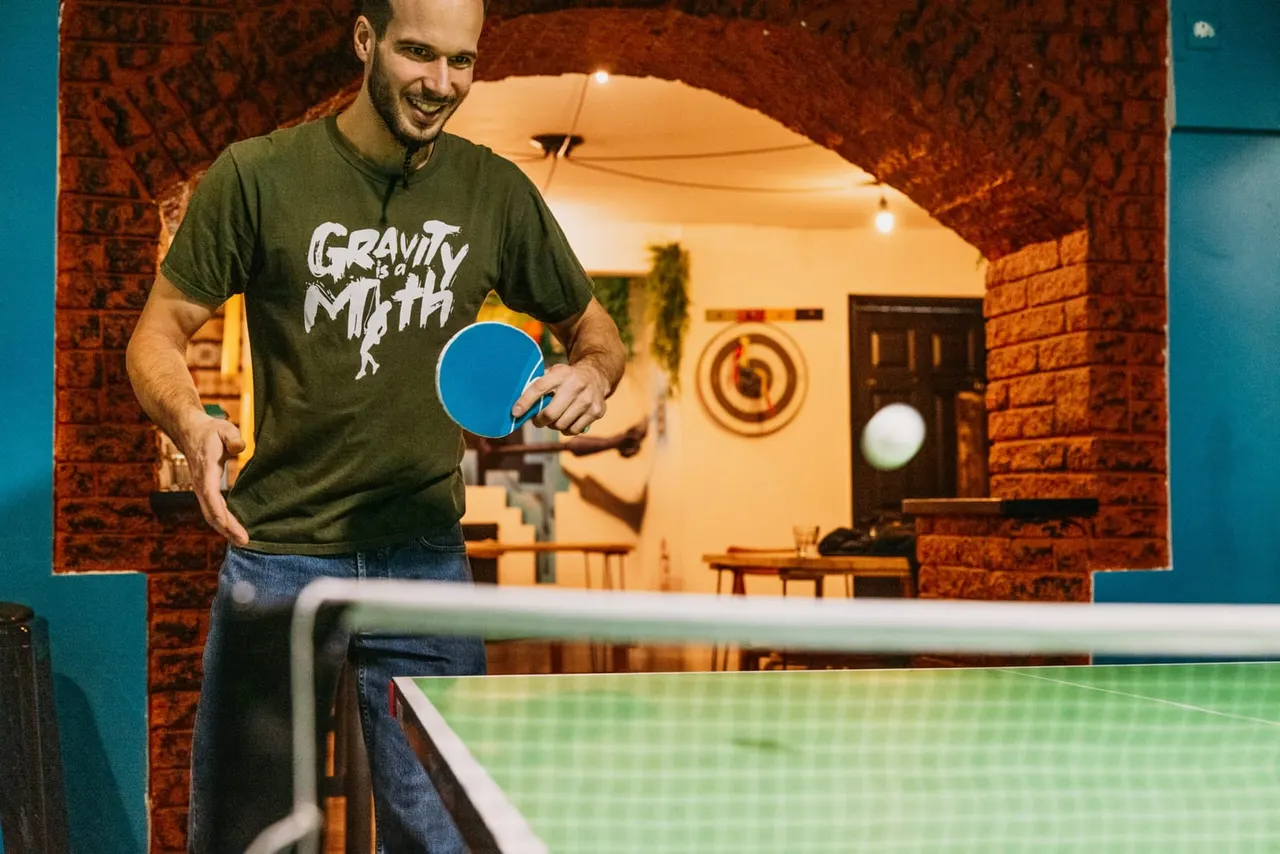 Man playing table tennis in a brick-walled games venue with a blue paddle and ball in motion.