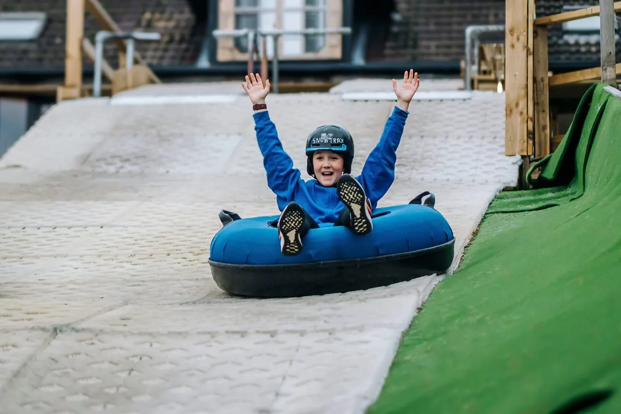 Child in blue jacket and helmet sledging down a slope on an inflatable tube with arms raised.