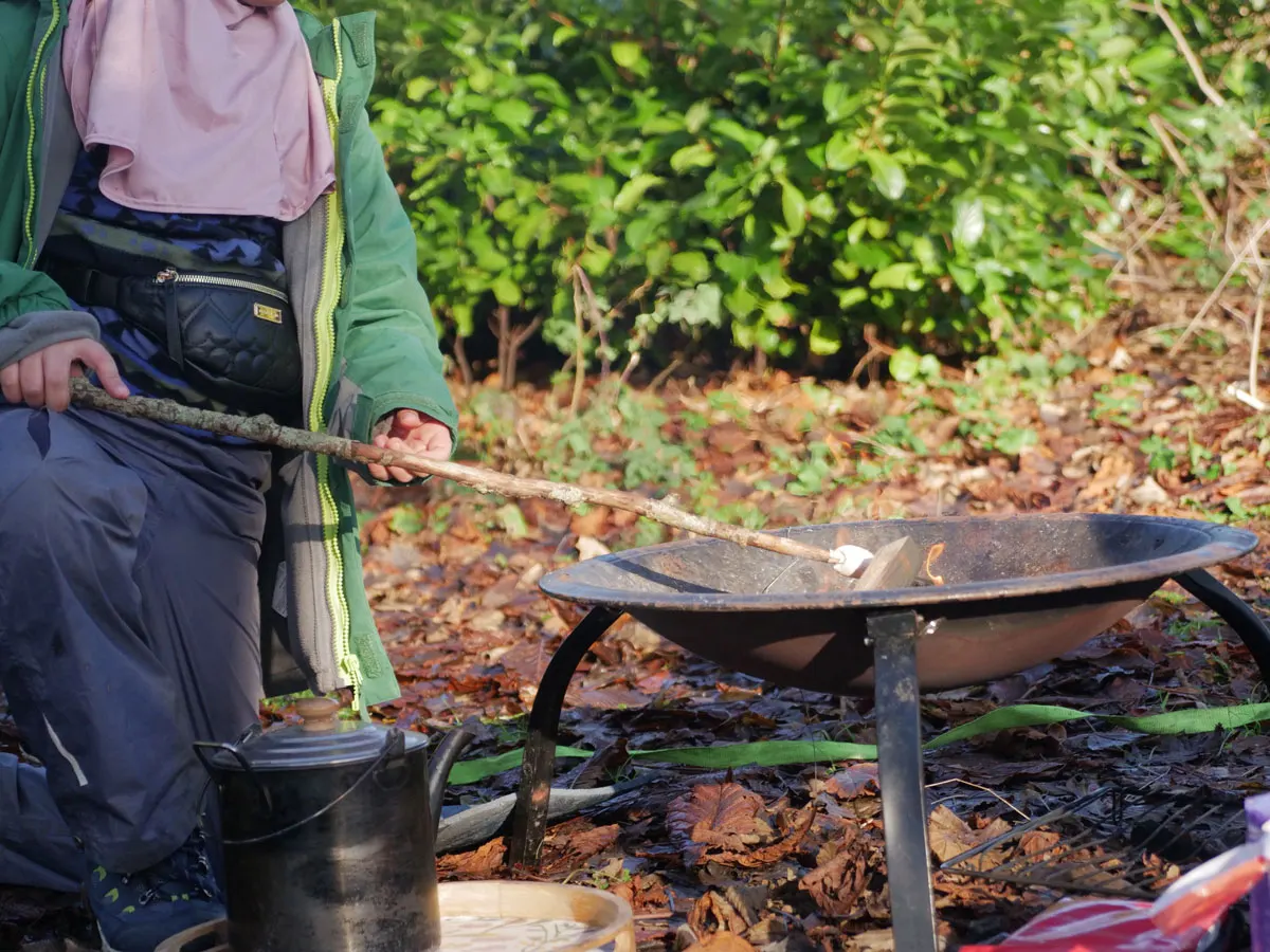 Child in green jacket tending a fire pit with a stick, surrounded by woodland and outdoor cooking equipment.