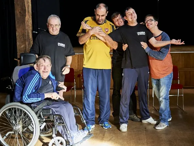 Group of six people posing together indoors, including a man in a wheelchair and others wearing casual clothing and branded shirts.